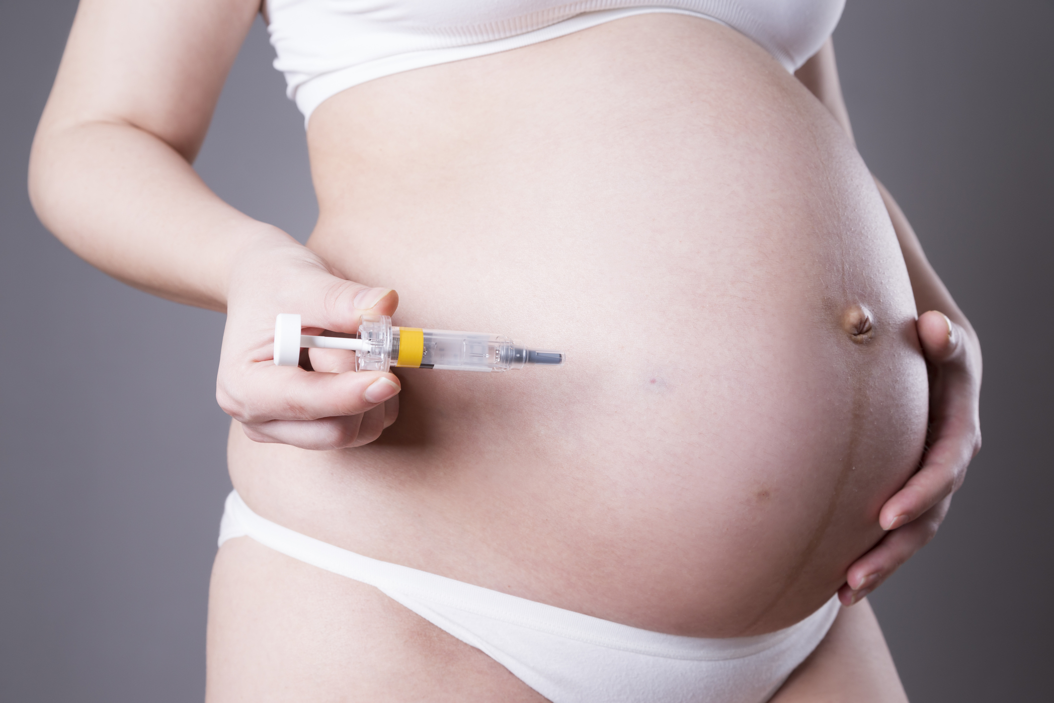 Young pregnant woman making injection in the stomach. Close-up studio shot on a gray background